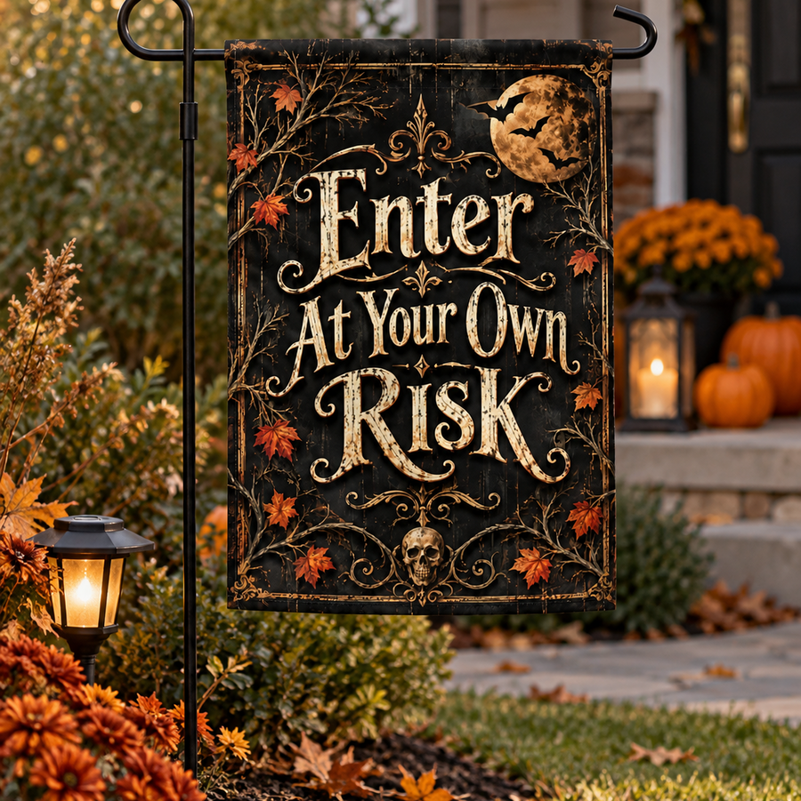 A decorative Halloween garden flag hanging from a black metal stand in a front yard. The flag has a dark background with ornate gold lettering that reads 'Enter At Your Own Risk,' plus a full moon, bats, skull, and autumn leaves.